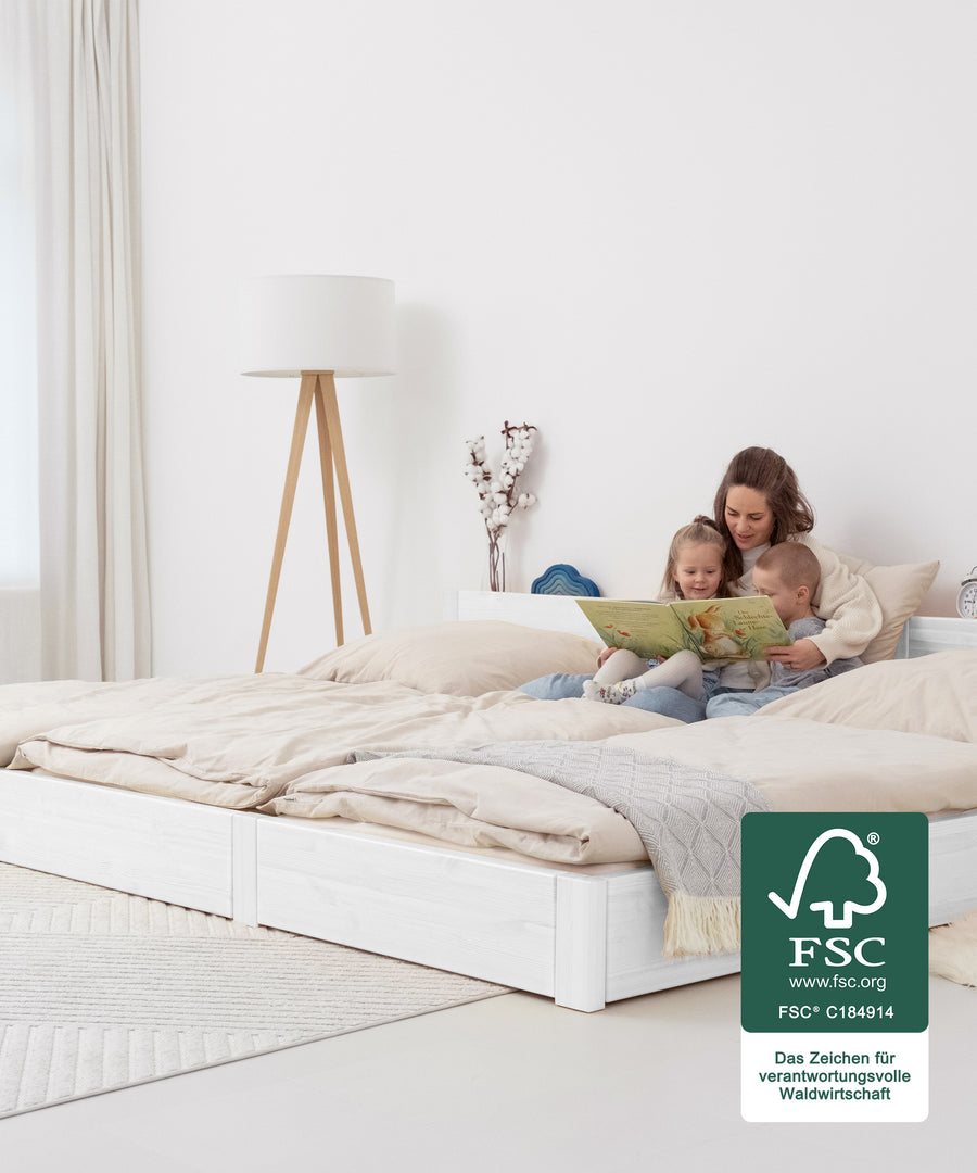 Das perfekte Familienbett*Für jedes Zuhause die passende Variante: Unser XXL Bett gibt es in zwei Höhen, beide erhältlich in weiß und transparent lackiert.|Familie sitzt auf Familienbett weiß bodentief und schaut ein Buch an.