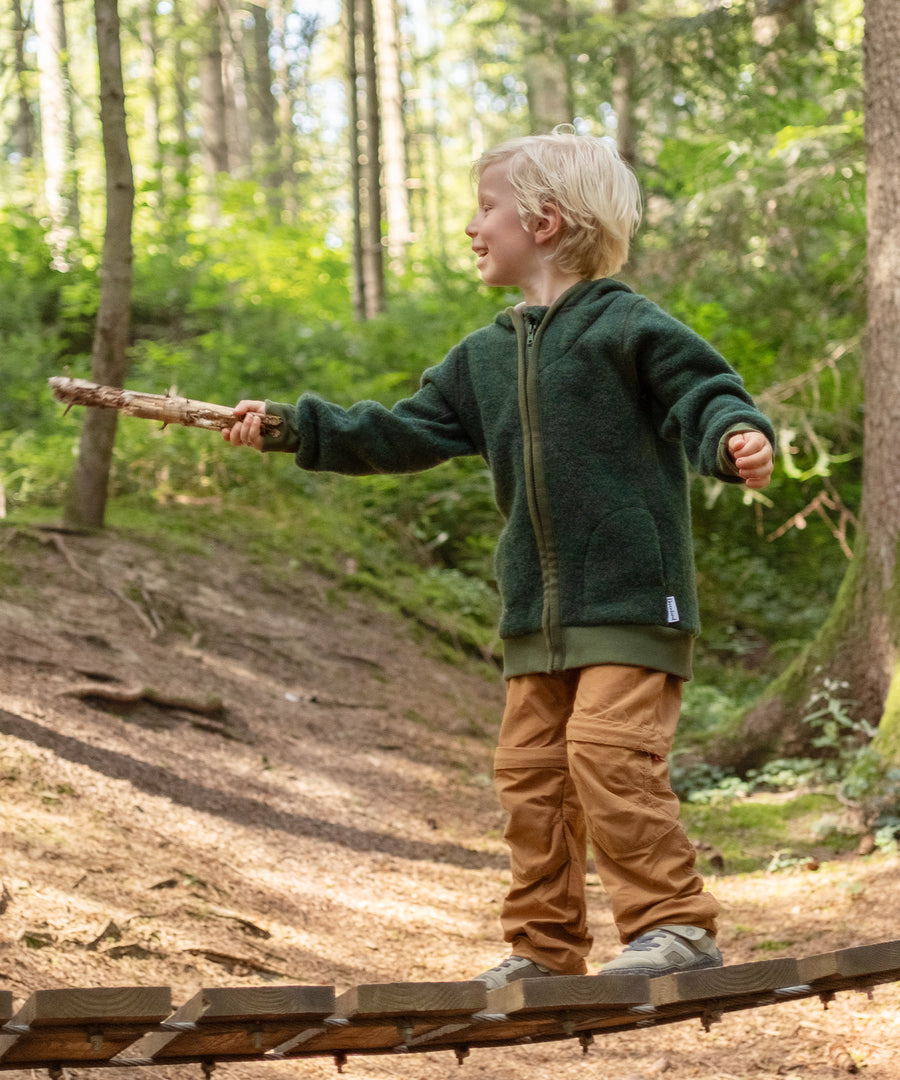 Beste Materialeigenschaften*Wollfleece ist atmungsaktiv, temperatur- und feuchtigkeitsregulierend. So bleibt dein Kind angenehm warm, ohne zu schwitzen.|Junge mit Wollfleece-Jacke Waldgrün spielt im Wald.
