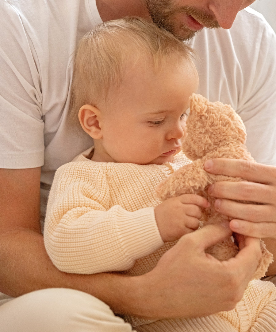 Baby sitzt bei seinem Vater auf dem Schoß, hält ein Kuscheltier und trägt einen kuscheligen Strickoverall aus Bio-Baumwolle.