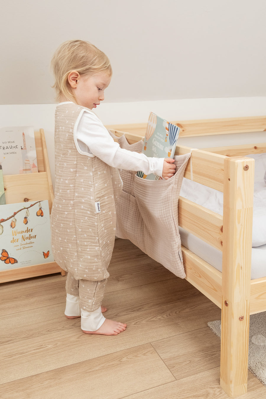Kind in Musselin-Sommerschlafsack mit Beinen steht vor Bett.