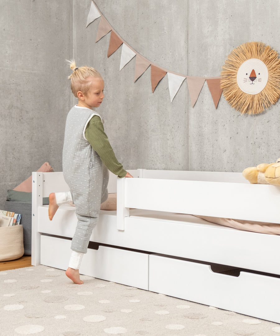 Abnehmbarer Rausfallschutz*Dank des Rausfallschutzes schlafen kleinere Kinder sicher. Er kann flexibel beidseitig montiert und leicht abgenommen werden.| Kind lehnt an Kinderbett Buche weiß.