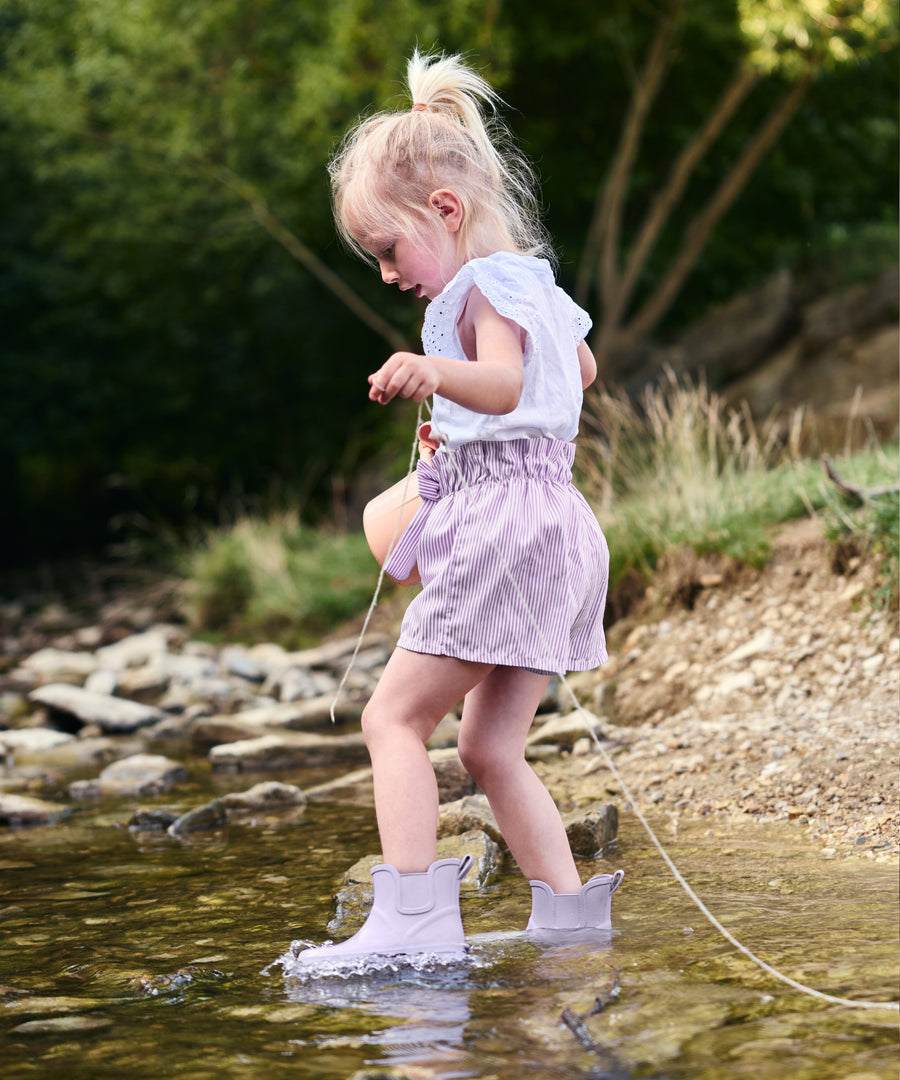 Unbeschwerter Pfützenspaß*In den Barfuß-Regenstiefeln bleiben kleine Füße sauber und trocken. Sie sind vollständig wasserdicht und sorgen für guten Halt.| Kind spielt im Wasser und trägt die Barfußgummistiefel in Flieder