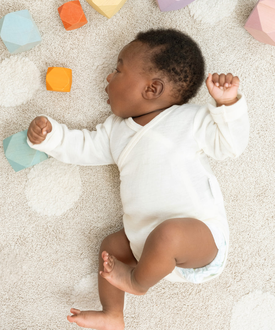 Temperaturausgleichend & sanft*Wie eine zweite Haut wärmt Merinowolle, wenn es kalt ist und kühlt, wenn es warm ist - der perfekte Begleiter zu jeder Jahreszeit.|Baby im Langarmbody liegt auf Teppich.